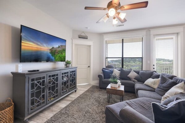 Una sala de estar con un televisor de pantalla plana grande, un juego de sofá gris, un ventilador de techo y vistas a través de las ventanas.