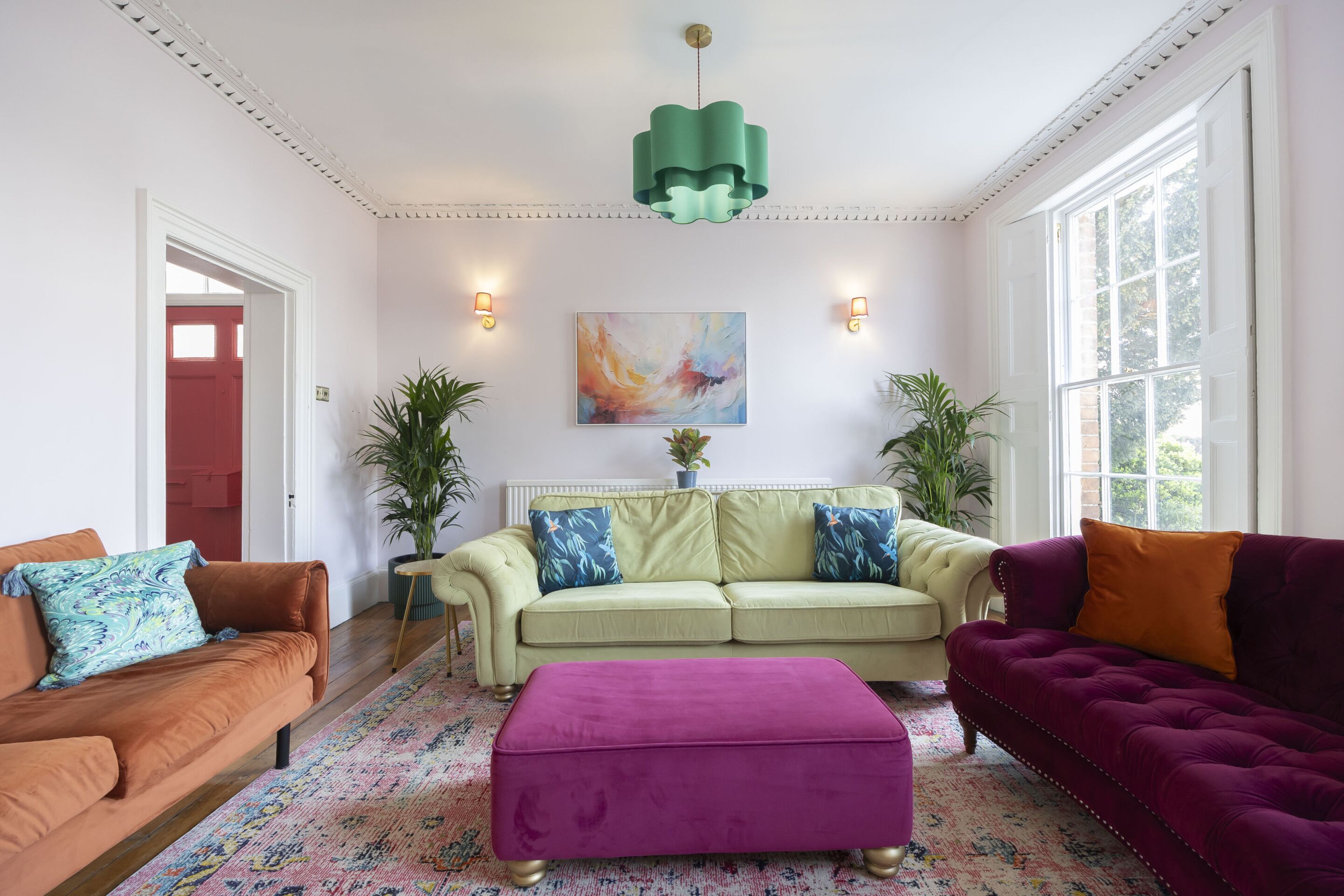 A living room with a green pendant light, a beige sofa with blue pillows, a purple sofa, and a pink ottoman.
