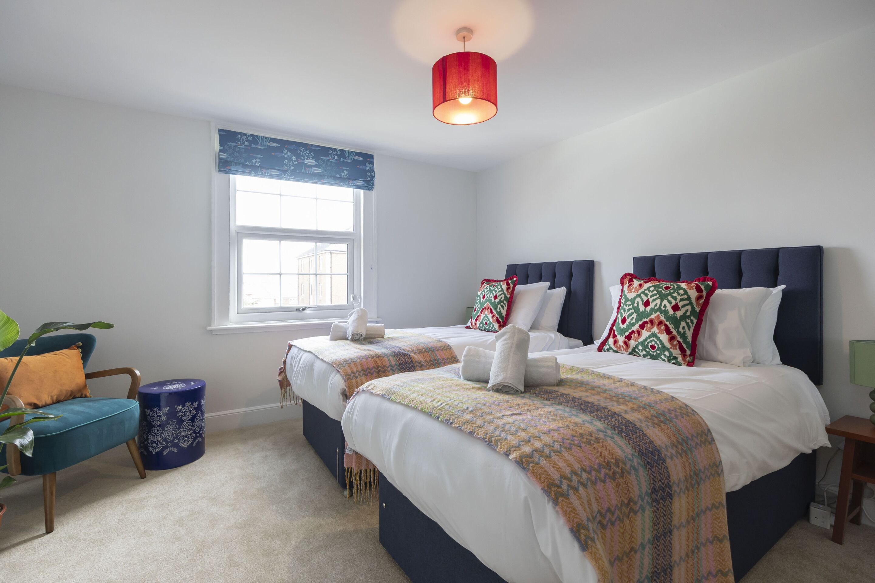 A bedroom with two beds, a window, a plant, and a lamp.