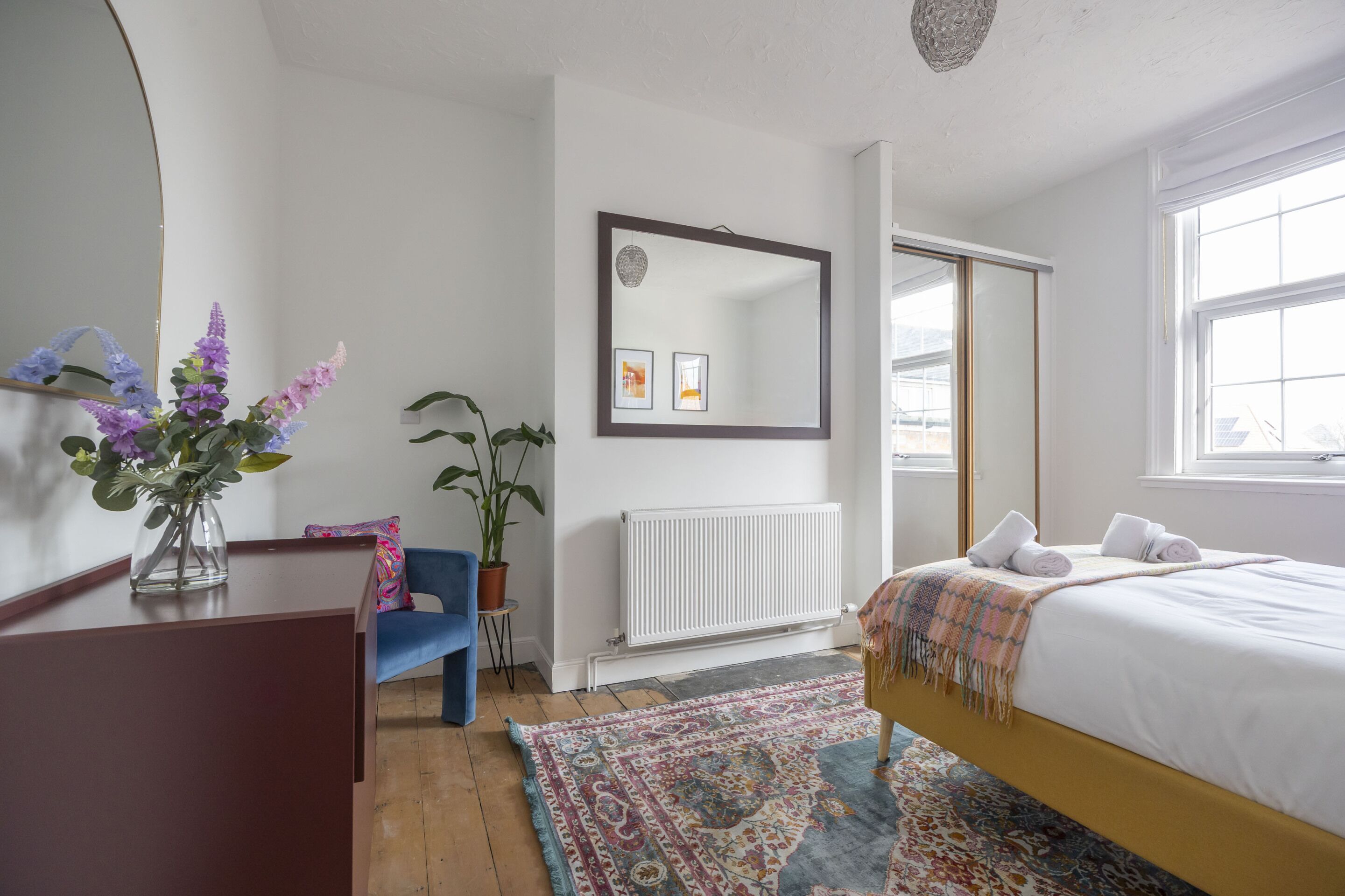 A bedroom with a bed, a dresser, a mirror, a radiator, a window, and a plant.