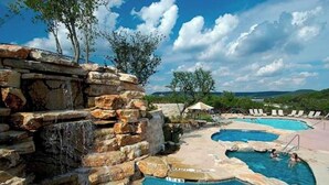 Ein Poolbereich mit Steinwänden und einem Wasserfall, umgeben von Liegestühlen und blauem Himmel.