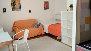 Una habitación con un escritorio blanco, una silla blanca, un sofá naranja, una mesita con una planta y una estantería blanca.