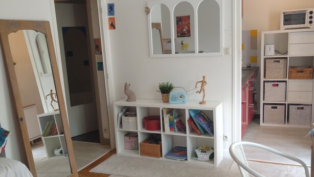 Una habitación con alfombra blanca, una silla blanca, una estantería blanca con cajas de almacenamiento, un espejo y un adorno colgante.