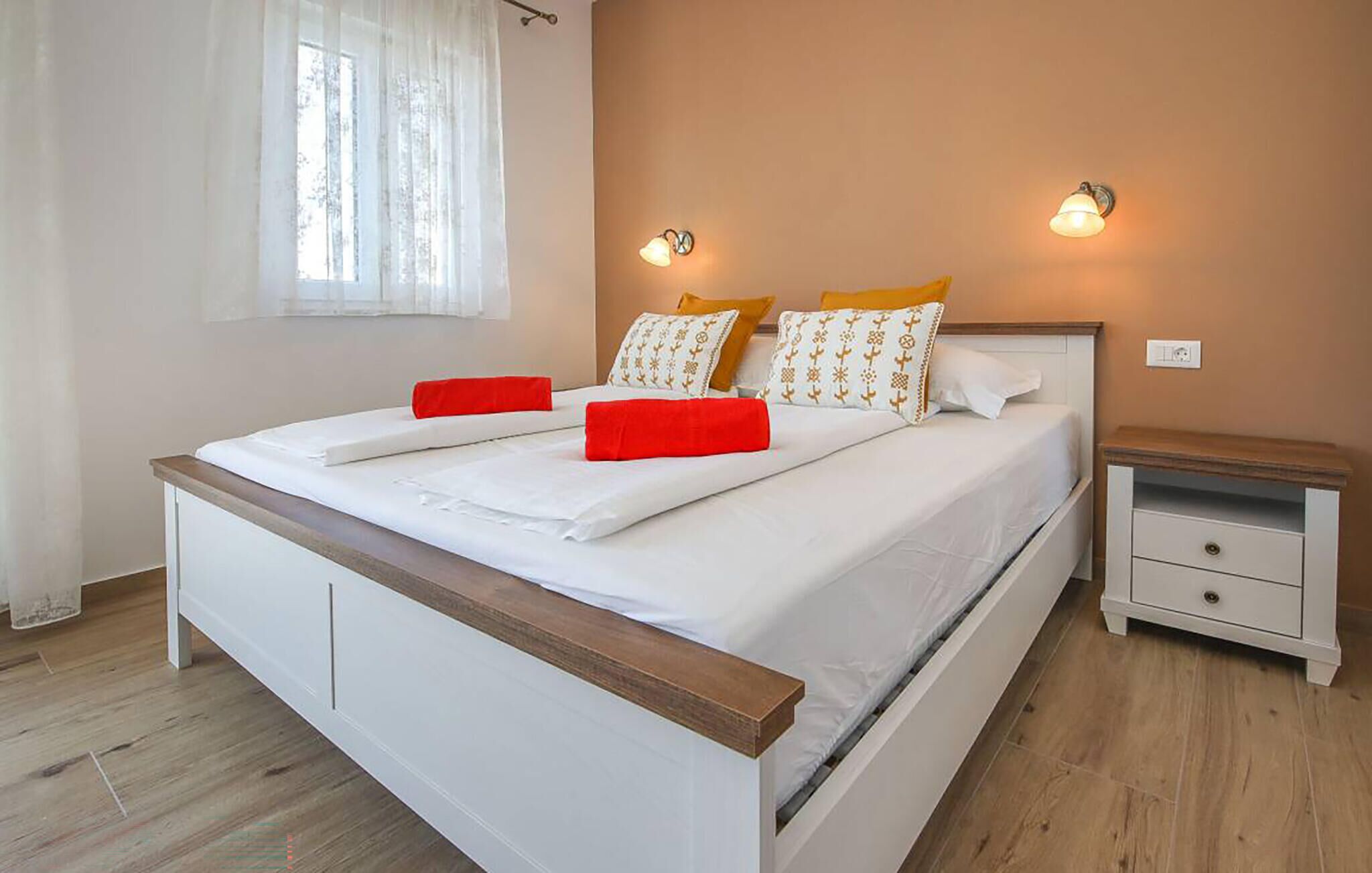 Una cama bien hecha con sábanas blancas y detalles rojos, una mesita de noche con cajón y una ventana con cortinas traslúcidas.