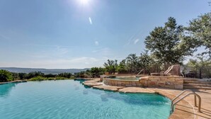 A clear blue infinity pool with a stone edge, surrounded by a landscaped area with trees and a view of distant hills.