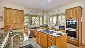 A spacious kitchen with wooden cabinets, a central island, stainless steel appliances, and large windows. - Hill Country Estate | 49 Acres | Pool | Game Room | Great Deals! (Lago Vista)