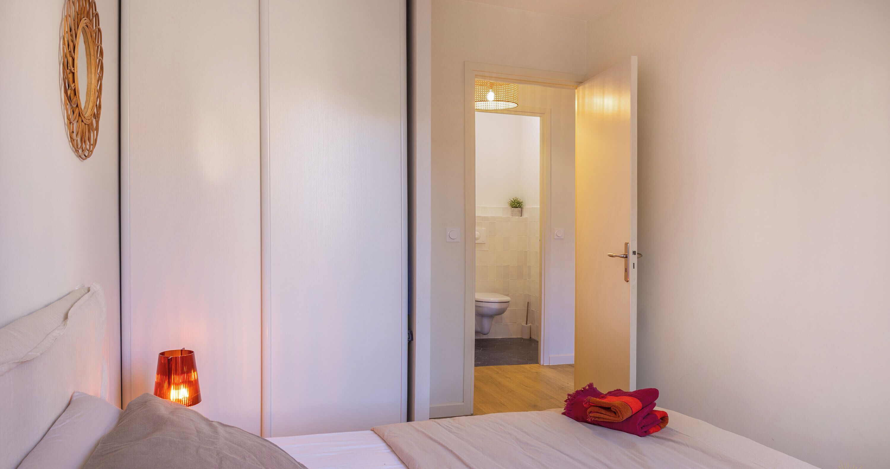 Un dormitorio con cama, una lámpara de noche, una puerta que da al baño y una planta pequeña en el mostrador del baño.
