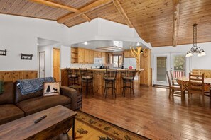 A spacious living area with a wooden ceiling, a sofa with a throw blanket, a coffee table, and a dining area with chairs and a table. - Leaf & Needle (Big Bear Lake)
