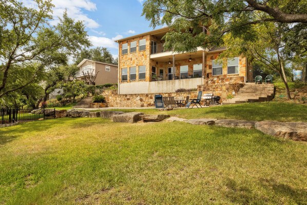 A two-story stone house with a balcony, a grassy yard, and a barbecue setup. - Lake Travis Waterfront Home: Boat Dock, Balcony, Winery | Amazing Views! (Marble Falls)
