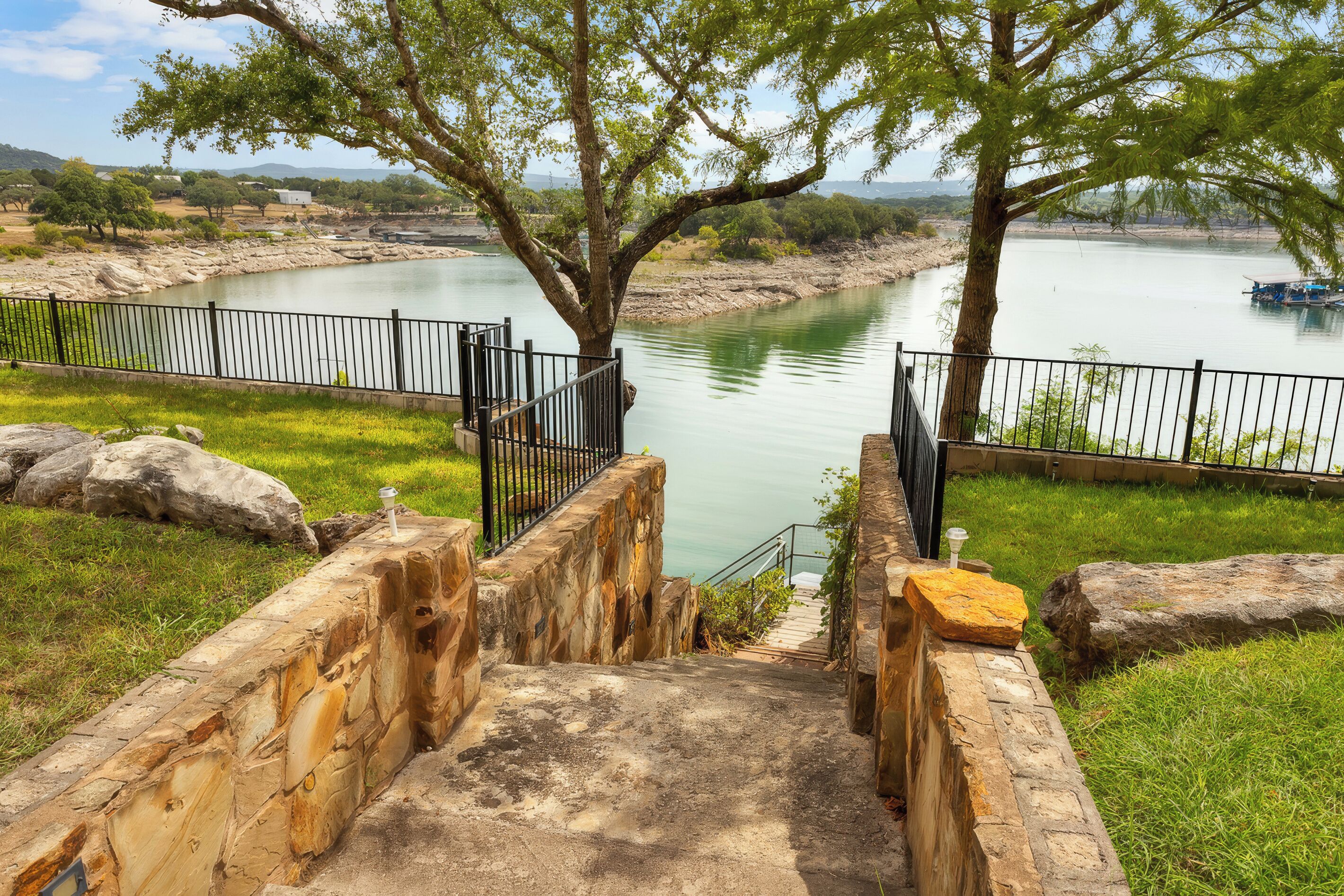 Una escalera de piedra que desciende hasta la orilla de un río, con una valla metálica y árboles.