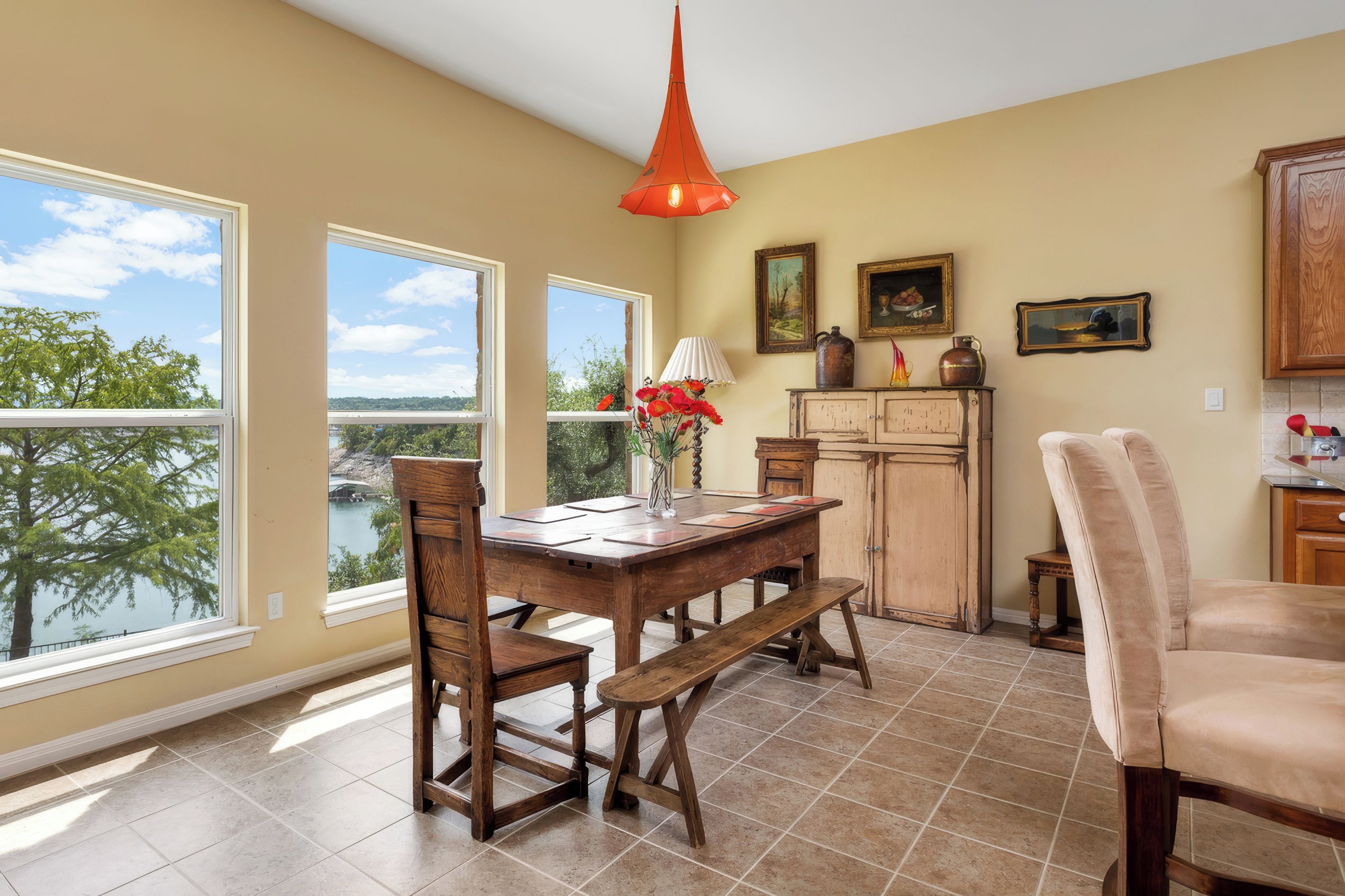 A dining area with a wooden table and chairs, a vase with red flowers, and a view of a lake through large windows.