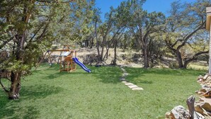 A backyard with a wooden play structure, a blue slide, and a stone pathway. - The Lazy Lakehouse - All LVPOA Amenities Available (Lago Vista)