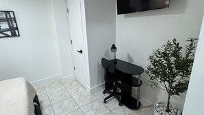 Ein Schlafzimmer mit einem Bett, einem Schreibtisch, einem Stuhl, einer Zimmerpflanze, einem Fernseher und einer Tür.