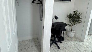 Ein Zimmer mit weißer Tür, einem Kleiderschrank mit Kleidung, einem Schreibtisch mit Stuhl, einer Topfpflanze und einem an der Wand montierten Fernseher.