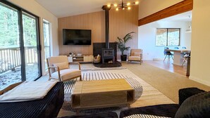 A modern living room with a fireplace, a flat-screen TV, and a dining area with a wooden table and chairs. - Secluded 5BR Cabin, Hot Tub, Views, Mt Hood (Rhododendron)