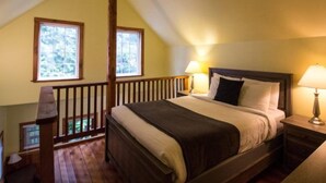 Un dormitorio con techo inclinado, estructura de cama de madera, dos lámparas de noche y una ventana con vista a árboles.