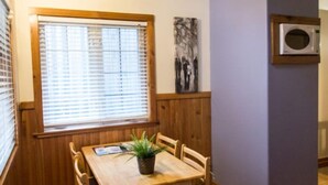A wooden dining table with chairs, a potted plant, a window with blinds, a wall-mounted microwave, and a framed artwork.