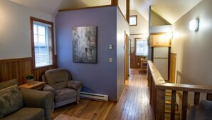 A loft-style living area with a high ceiling, wooden flooring, a large painting on the wall, and a view of the kitchen through an open door.