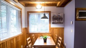 Un comedor con mesa y sillas de madera, una planta en maceta, una lámpara colgante y un ventanal con persianas.
