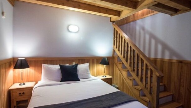 Un dormitorio con techo de madera, cama con ropa de cama blanca y negra, dos lámparas de noche y una escalera de madera.