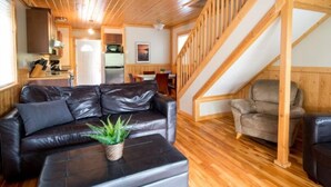 A cozy cabin interior with a wooden staircase, a leather sofa, a dining area, and a kitchen.