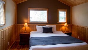 A bedroom with a sloped ceiling, two windows, a bed with white bedding and a dark throw, and two bedside lamps.