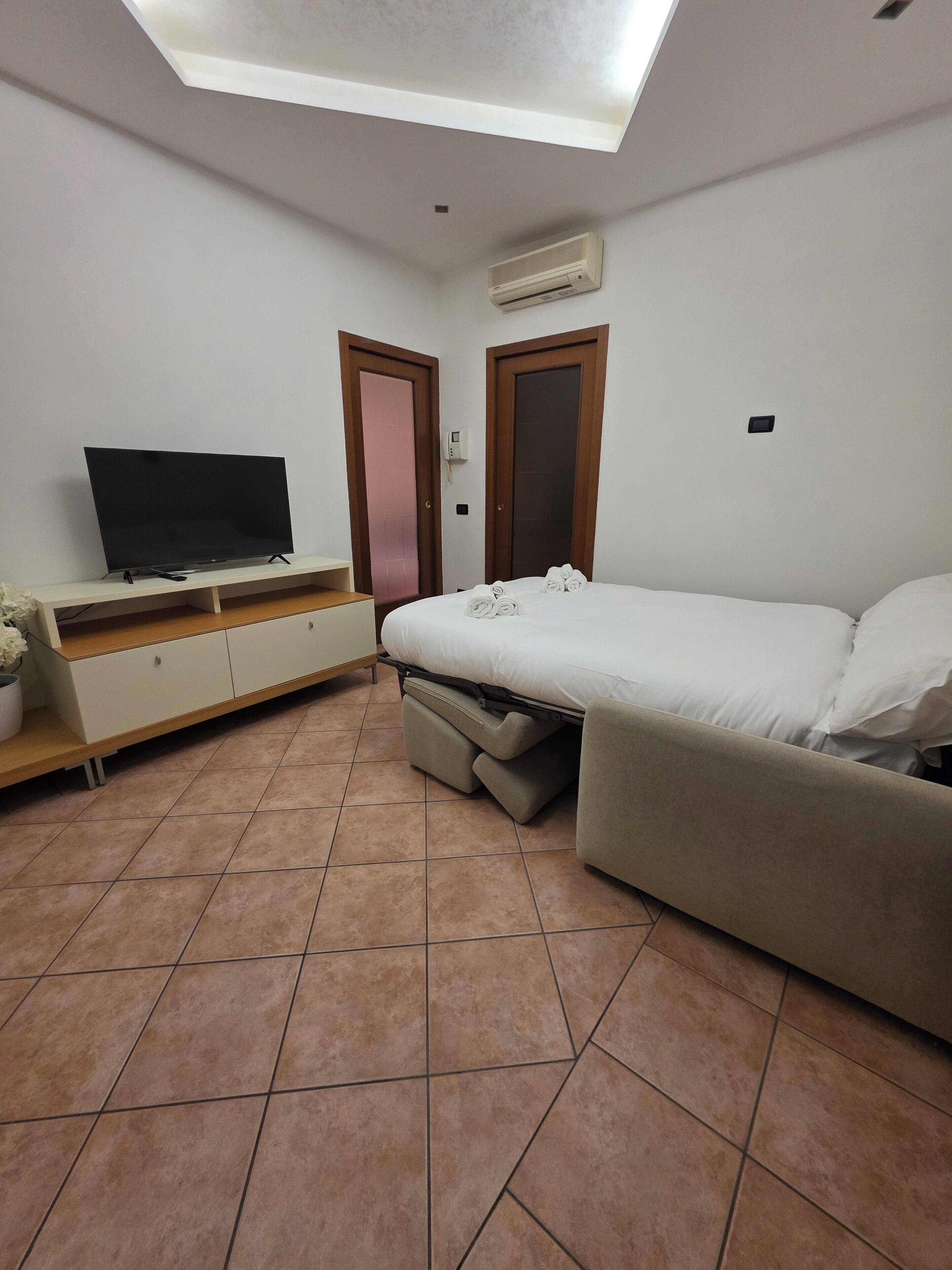 A bedroom with a bed, a TV on a stand, a ceiling light, and a wall-mounted air conditioner.