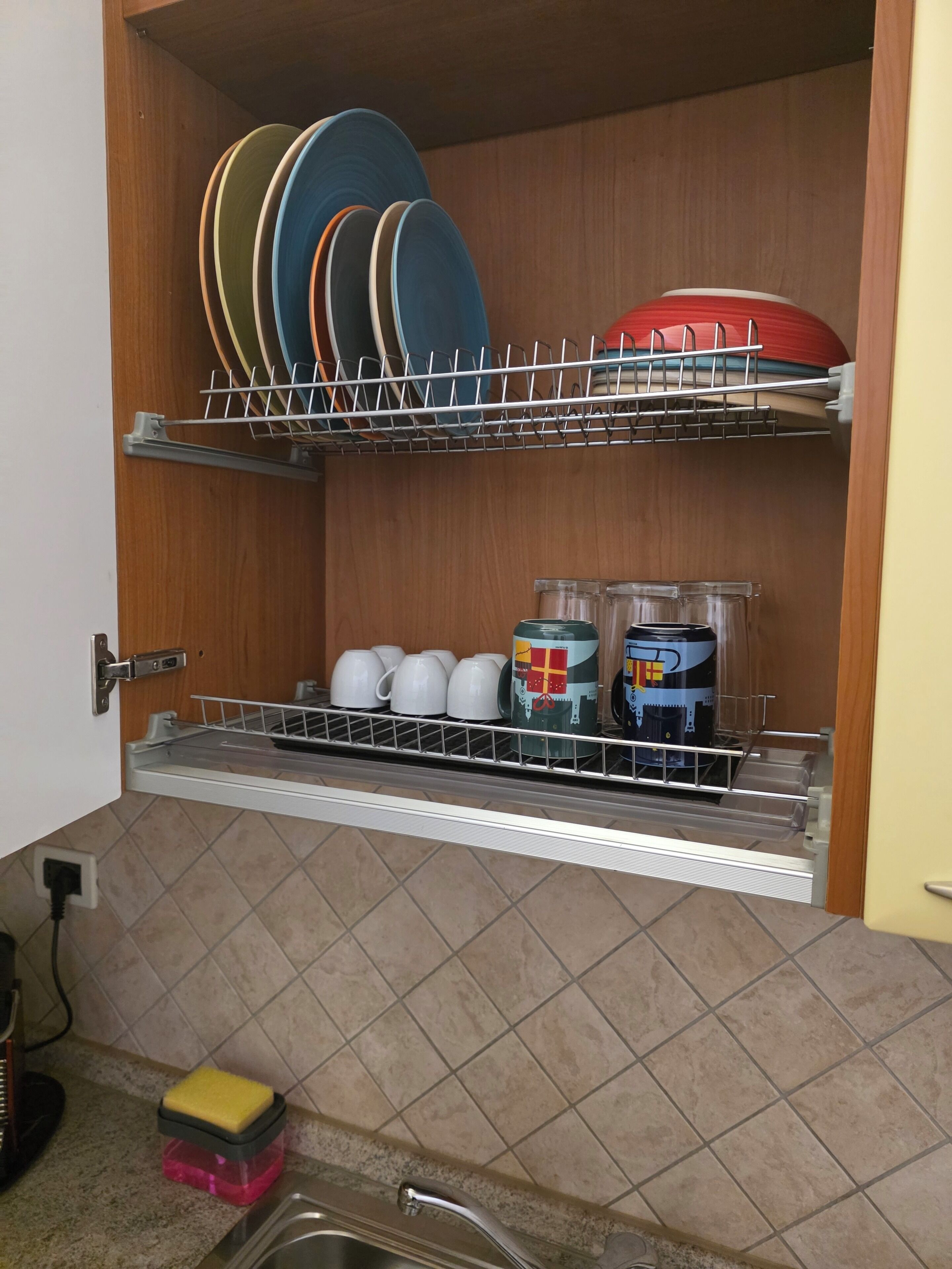 A kitchen with a dish rack, a shelf with plates, and a countertop with a sponge and containers.