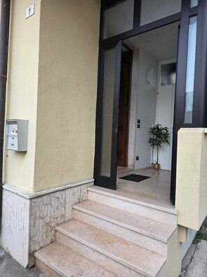 Entrada de um edifício com uma escadaria que leva a porta, um vaso de planta e um quadro de eletricidade na parede.