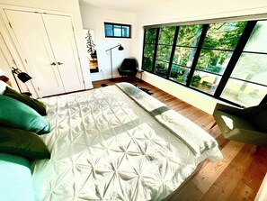 A bedroom with a large bed, a view of greenery through a window, a desk, and a chair.