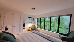 A bedroom with a large window, a bed, a chair, and a desk with a lamp.