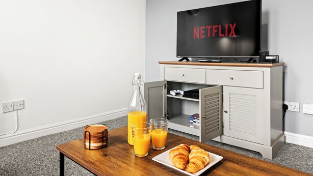 Een kamer met een televisie waarop 'NETFLIX' staat, een houten tafel met een fles sinaasappelsap, glazen en croissants, en een klein kastje met een magnetron en andere spullen.