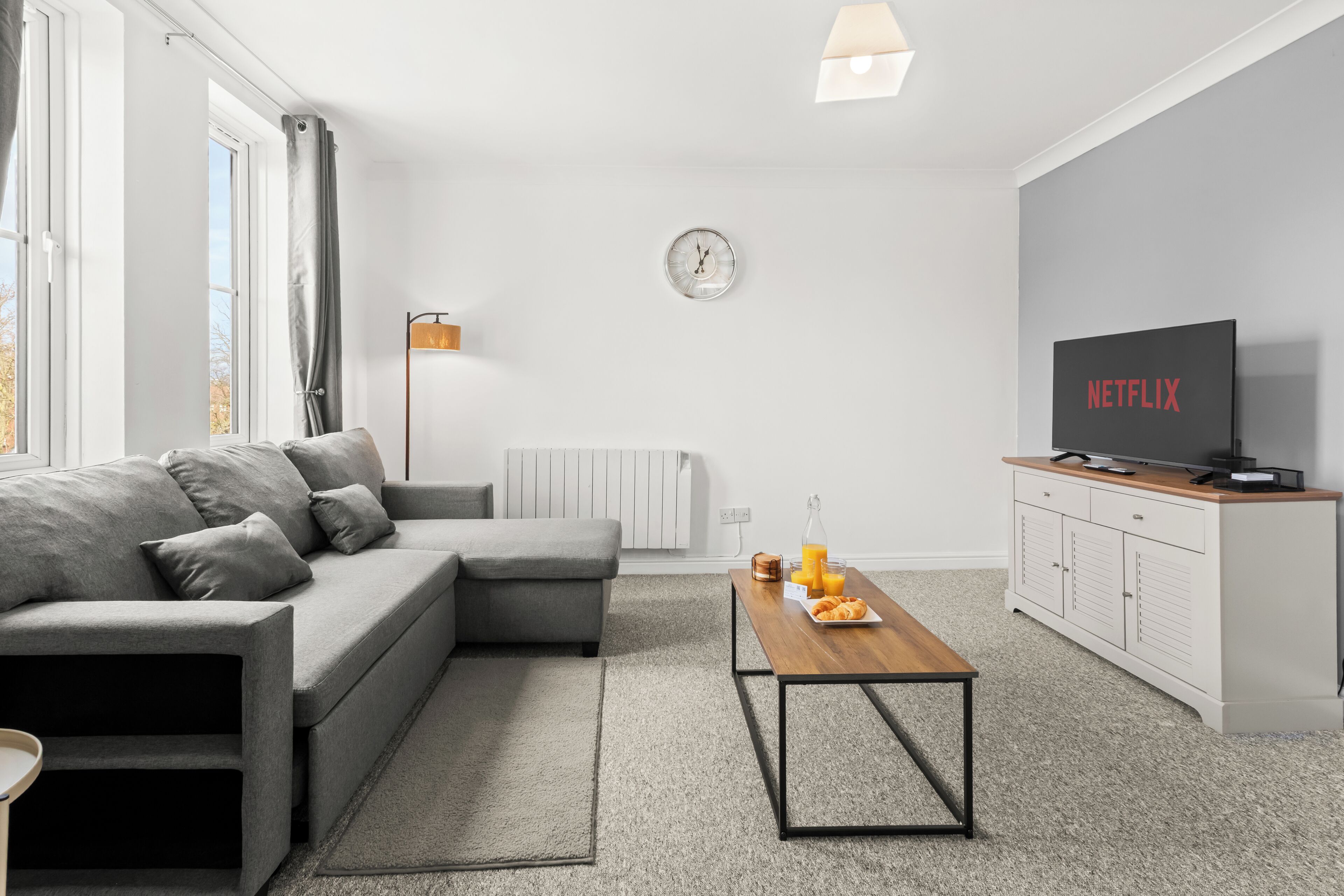 A modern living room with a grey sectional sofa, a wooden coffee table, a TV on a white cabinet, and a radiator.