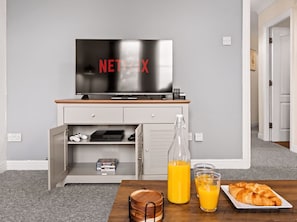 Ein Zimmer mit Fernseher, der 'NETFLIX' anzeigt, einem Holztisch mit Orangensaft, Gläsern und einem Croissant sowie einem weißen Schrank mit Schubladen.
