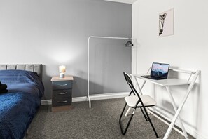 Un dormitorio con cama, escritorio con ordenador portátil, silla y mesita de noche con lámpara.