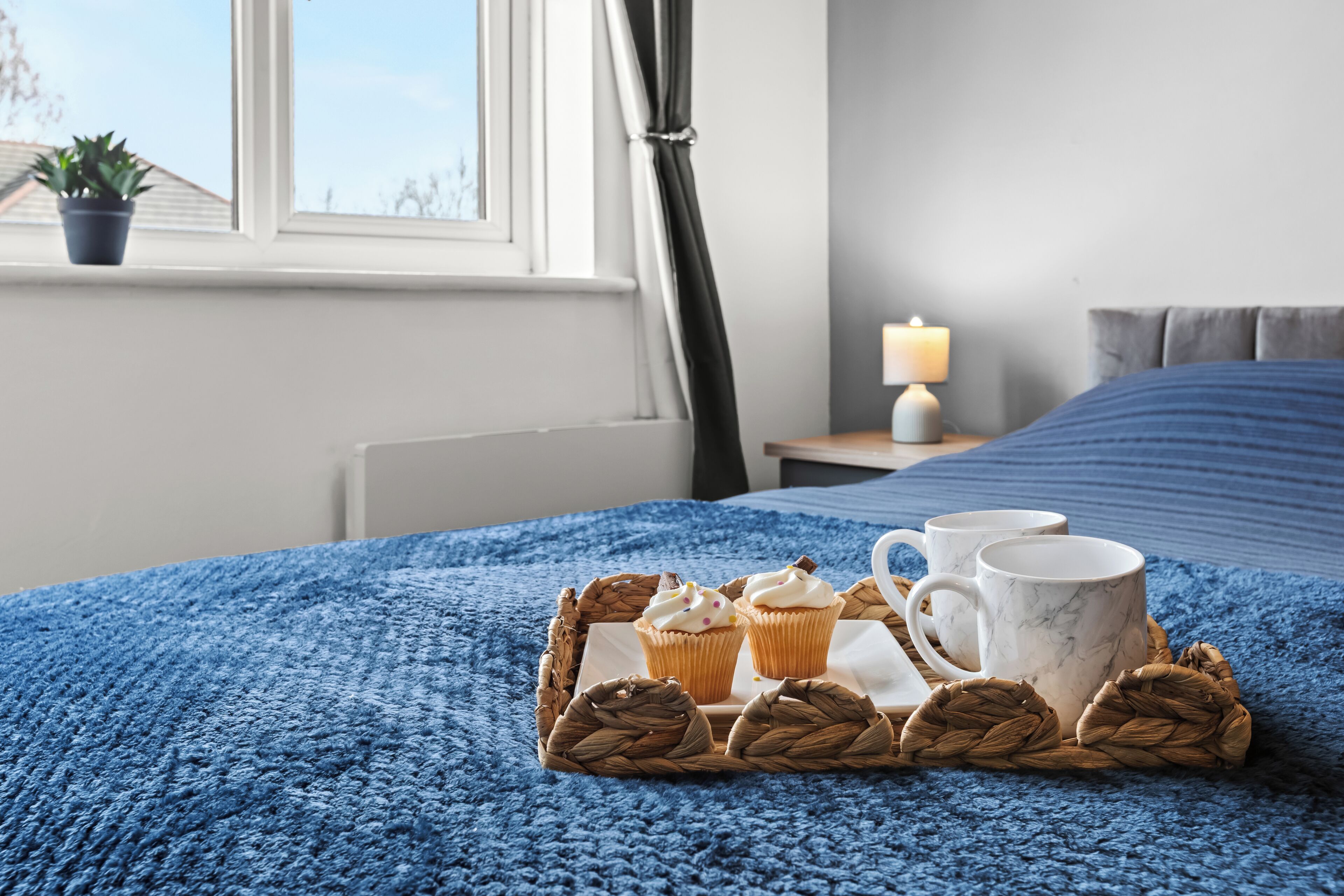 A neatly made bed with a blue blanket, a bedside table with a lamp, and a tray with cupcakes and mugs on a blue carpet.