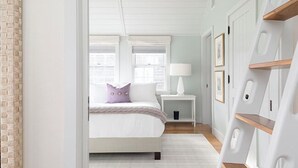 A bedroom with a bed, nightstand, and bedside lamp. There are windows with blinds, a brick wall, and a white door with a wooden shelf. - 2.5 bedroom, 2.5 bathroom (Nantucket)