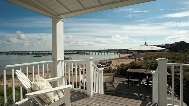 A covered deck with white railings, a bench, and a table with chairs, overlooking a beach and a pier. - 1 Bedroom, 1 bathroom (Nantucket)