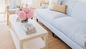 A living room with a light blue sofa, a white coffee table with books and flowers, a wicker chair, a round mirror, and a lamp. - 2 bedroom, 2.5 bathroom (Nantucket)