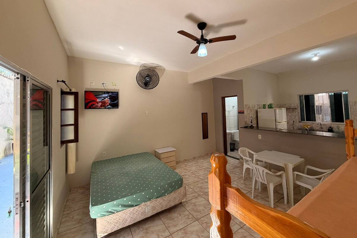 A room with a bed, a ceiling fan, a TV, a window, and a kitchenette.