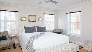 A bedroom with a large bed, two bedside tables with lamps, a ceiling fan, and two windows with curtains.
