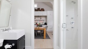 A modern bathroom with a shower, a sink, and a mirror. There is a dining area visible through an open door. - 1 Bedroom with loft, 1 bathroom (Nantucket)