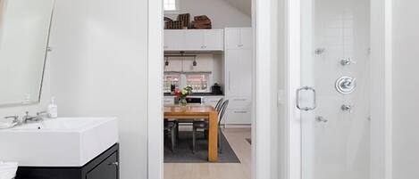 Un baño moderno con ducha, lavamanos y espejo. Se ve un comedor a través de una puerta abierta.