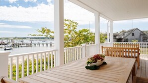 A covered outdoor area with a wooden table and chairs, a white railing, and a view of boats and buildings across the water. - 5 bedroom, 7 bathroom (Nantucket)