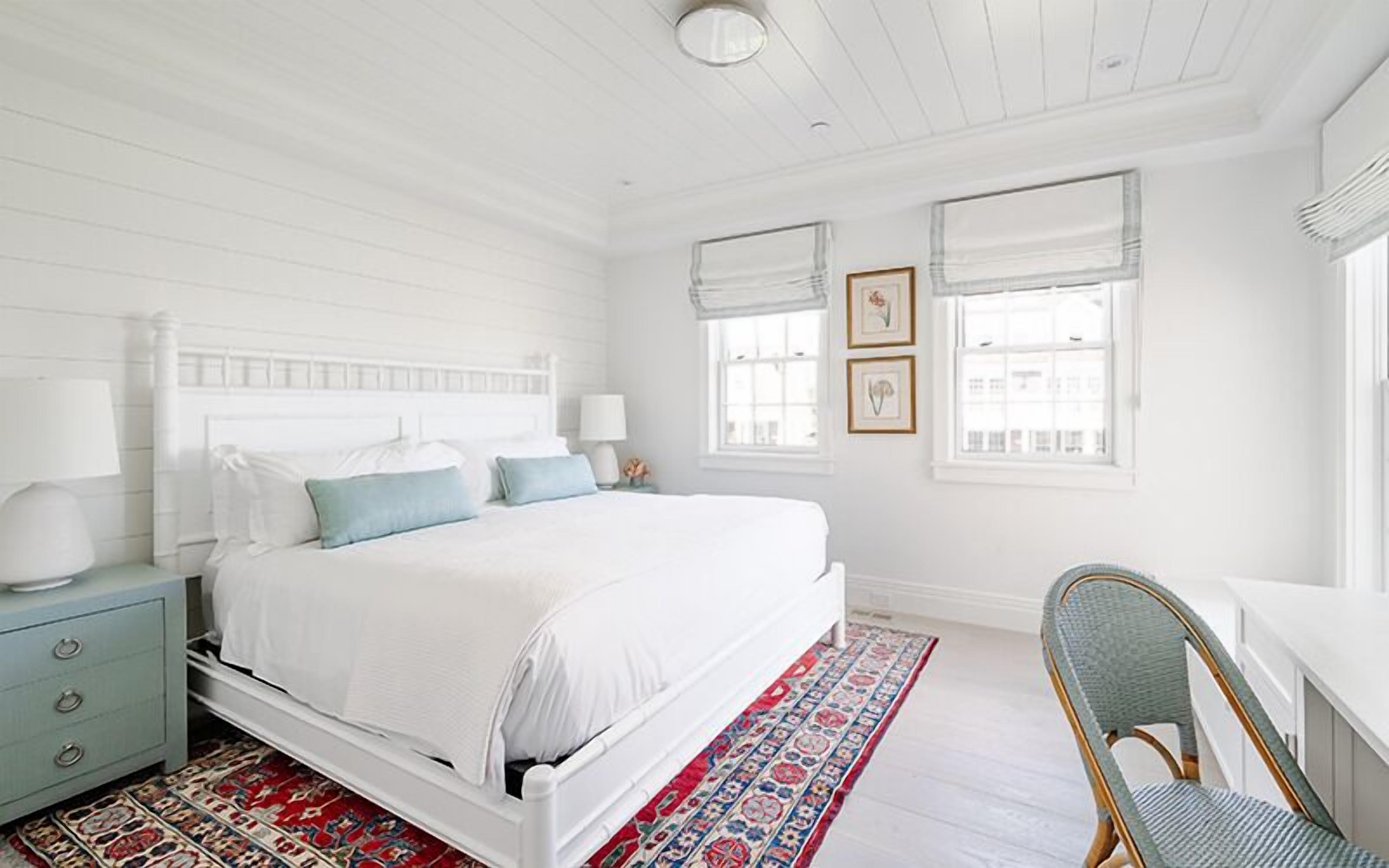 Un dormitorio blanco con cama, mesitas de noche, una silla y una alfombra estampada.