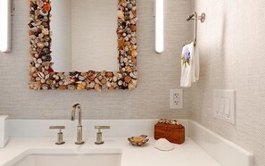 Een badkamer met een wastafel, drie kranen, een spiegel met een schelpenrand, een handdoekhouder en een klein houten doosje met schelpen.