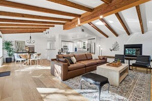 Ein großzügiger Wohnbereich mit freiliegenden Holzbalken, einem braunen Ledersofa, einem Couchtisch und einem Essbereich mit Pflanze und Kunstwerk.