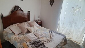 Een opgemaakt bed met een bedsprei en kussens, een nachtkastje met een lamp, en een raam met een dunne gordijn.