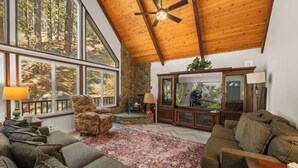A spacious living room with a large window, a fireplace, and a ceiling fan. - Eads by the Lake (Fawnskin)