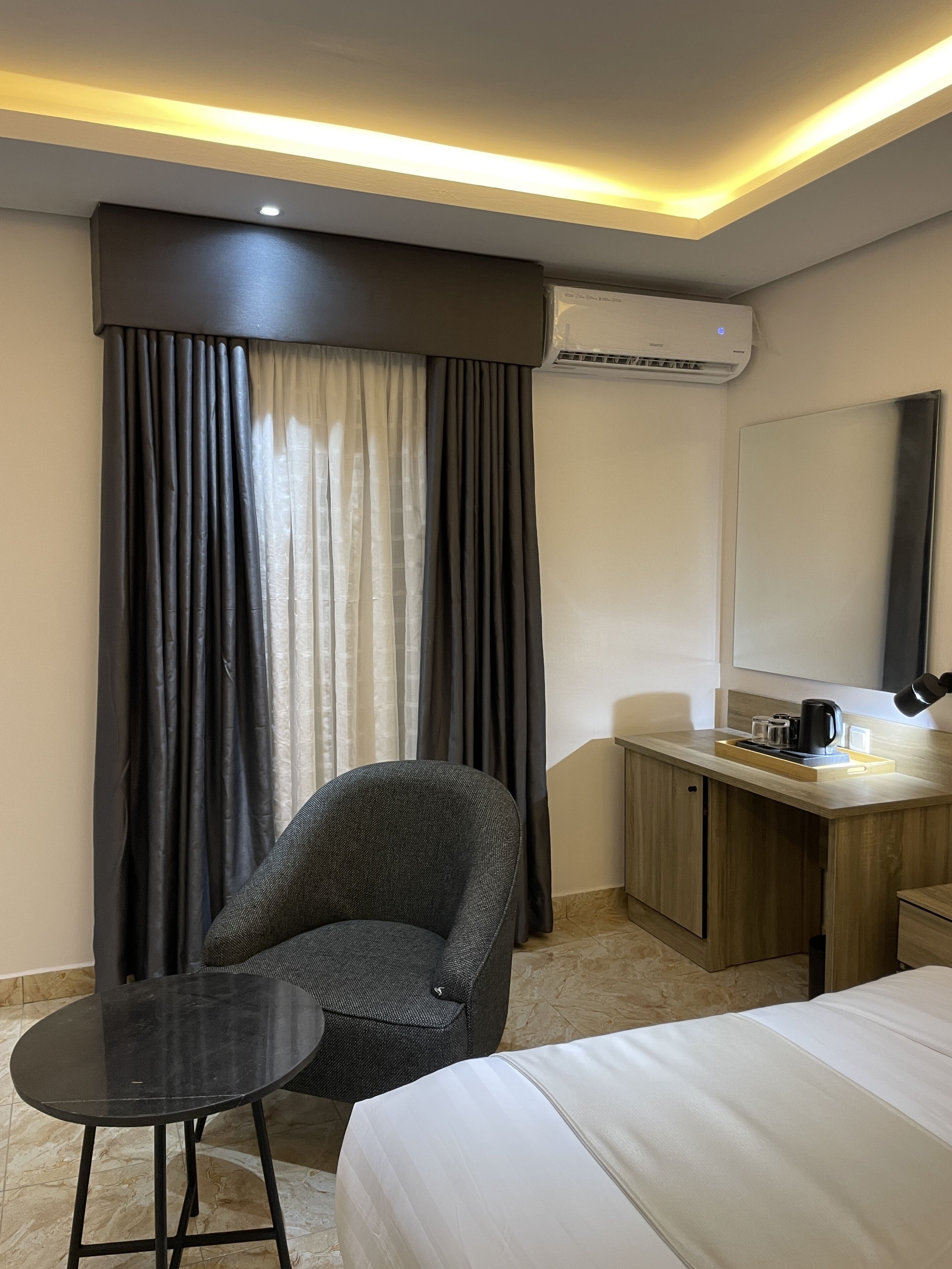 A hotel room with a bed, a desk, a chair, a small table, an air conditioner, and a window with curtains.